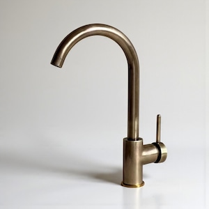 Peut inclure: Un robinet de cuisine en bronze avec un bec courbé et une poignée à levier unique. Le robinet a un design moderne et est posé sur un fond blanc. La base du robinet a un anneau de couleur or.