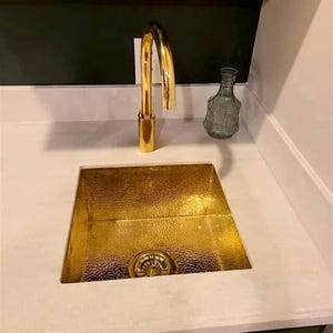 Può includere: Un lavello da cucina dorato con un rubinetto abbinato. Il lavello è rettangolare e ha una superficie strutturata. Una bottiglia di vetro decorativa è appoggiata sul piano di lavoro. Il rubinetto ha un beccuccio curvo.