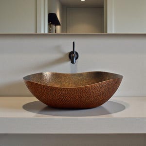 K&ouml;nnte beinhalten: Ein geh&auml;mmertes Kupferwaschbecken mit gewelltem Rand. Das Waschbecken hat eine dunkelbraune Oberfl&auml;che und ist auf einer hellgrauen Arbeitsplatte montiert. Ein schwarzer Wasserhahn ist an der Wand &uuml;ber dem Waschbecken angebracht.