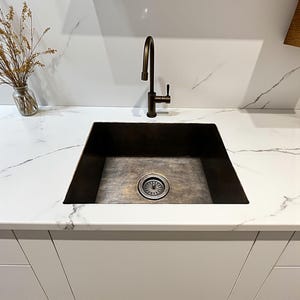 May include: A dark bronze-colored, square kitchen sink with a matching faucet. The sink is set into a white countertop with gray veining. A glass vase with dried plants sits to the left of the sink.
