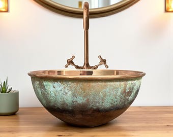Könnte beinhalten: Ein kupferfarbenes Waschbecken mit grüner Patina und einer bronzefarbenen Armatur. Das Waschbecken hat eine runde Form und steht auf einer Holzoberfläche. Die Armatur hat zwei kreuzförmige Griffe.