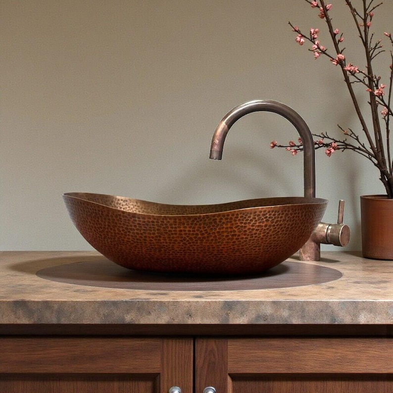 K&ouml;nnte beinhalten: Ein geh&auml;mmertes Kupferwaschbecken mit geschwungener Armatur. Das Waschbecken ist oval und steht auf einer Holzplatte. Eine kleine Pflanze mit rosa Bl&uuml;ten befindet sich im Hintergrund. Armatur und Waschbecken haben eine passende Kupferoberfl&auml;che.