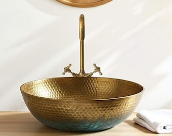May include: A hammered brass vessel sink with a vintage faucet. The sink has a round shape with a textured surface and a two-tone finish, transitioning from gold to teal. A white folded towel rests on the wooden surface.
