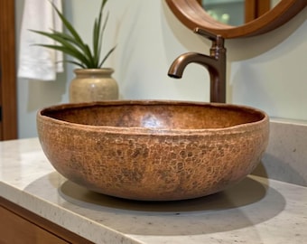 May include: A hammered copper vessel sink with a textured surface. The sink is oval-shaped and sits on a light-colored countertop. A bronze faucet and a round mirror are visible in the background, suggesting a bathroom setting.
