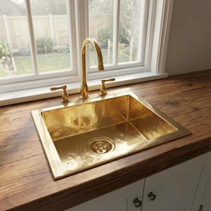 Peut inclure: Un évier de cuisine rectangulaire en laiton avec une finition polie et un robinet en laiton à arc élevé assorti. L'évier dispose d'une bonde centrale et de deux poignées à levier séparées pour le contrôle de l'eau, encastré dans un plan de travail en bois.