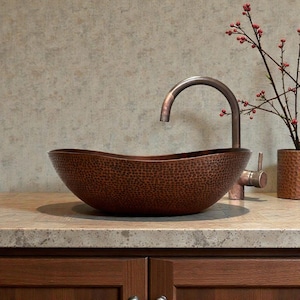 K&ouml;nnte beinhalten: Ein geh&auml;mmertes Kupferwaschbecken mit geschwungener Armatur. Das ovale Waschbecken steht auf einer Arbeitsplatte &uuml;ber dunklen Holzschr&auml;nken. Rechts vom Waschbecken steht eine kleine Vase mit roten Bl&uuml;ten.