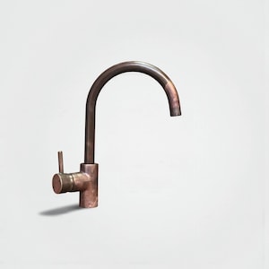 May include: A bronze-colored kitchen faucet with a curved spout and a single-handle lever. The faucet has a cylindrical base and a simple, modern design. The finish has a slightly aged appearance.