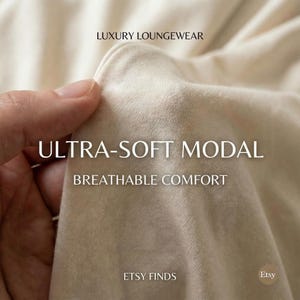 May include: Close-up of a beige fabric with the text "ULTRA-SOFT MODAL" and "BREATHABLE COMFORT". The words "LUXURY LOUNGEWEAR" and "ETSY FINDS" are also visible. The fabric is being held by a hand.