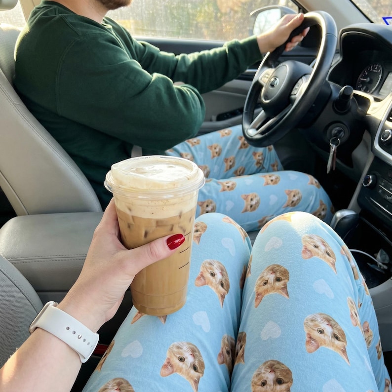 May include: A person holding a Starbucks iced coffee in a clear plastic cup with a white lid. The person is wearing a white watch and light blue pajama pants with a repeating pattern of cat faces and white clouds.
