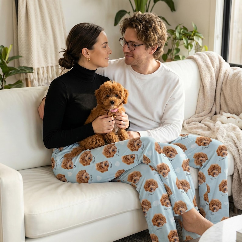 May include: A couple sits on a white couch, holding a small brown dog. The person's legs are covered by light blue pajama pants patterned with dog faces and the word "Friend". The dog has a pink collar.