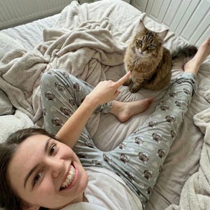 May include: A brown tabby cat sits on a bed next to a person wearing light gray pajama pants with a cat pattern. The person is pointing at the cat. The bed has a light gray comforter and a beige blanket.