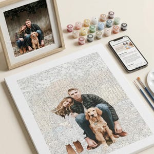 May include: A paint-by-numbers canvas depicting a couple and a dog, alongside a framed photograph of the same image. Includes paint pots, a smartphone with instructions, and paintbrushes. The canvas is pre-printed with numbered sections for painting.
