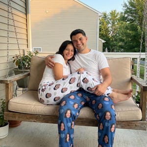 May include: A couple sits on a wooden swing, wearing matching white t-shirts. One person wears white pajama pants printed with faces, while the other wears blue pajama pants with faces and blue hearts. The swing has a beige cushion.