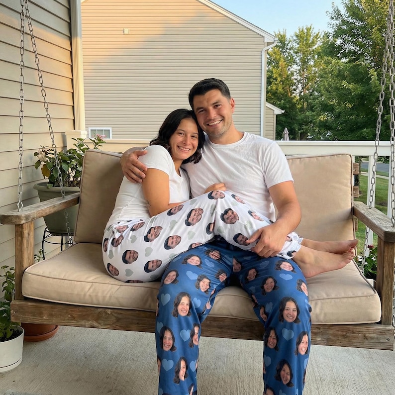 May include: A couple sits on a wooden swing, wearing matching white t-shirts. One person wears white pajama pants printed with faces, while the other wears blue pajama pants with faces and blue hearts. The swing has a beige cushion.