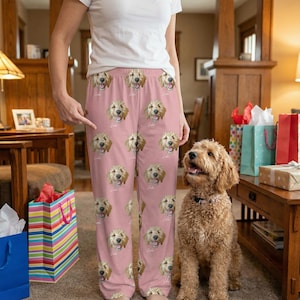 May include: Pink pajama pants featuring a repeating pattern of smiling golden doodle faces. The pants are loose-fitting and have a comfortable, casual style. A golden doodle dog sits nearby. The pajama pants are a unique and personalized gift.