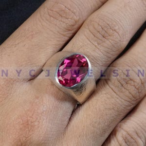May include: A silver ring with a large, oval-cut, magenta gemstone. The ring has a thick band and a simple, modern design. The gemstone is the focal point, catching the light and displaying a vibrant color.