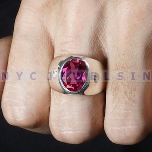 Puede incluir: Un anillo de plata con una gema grande, de corte ovalado, de color rosa intenso. El anillo tiene una banda ancha y pequeñas piedras de acento a cada lado de la piedra principal. El anillo está en una mano.