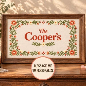 Puede incluir: Un bordado de punto de cruz rectangular en un marco de madera oscura con el texto The Cooper's en letras rojas, rodeado por un borde decorativo de hojas verdes y pequeñas flores naranjas sobre un fondo de tela blanca.
