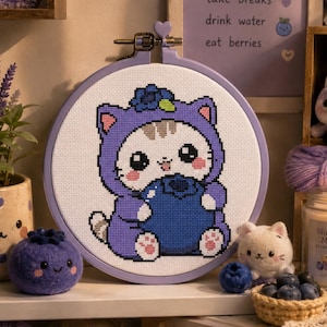 Könnte beinhalten: Ein runder Stickrahmen mit einem Kreuzstichmuster eines weißen Kätzchens, das eine violette Kapuze mit Katzenohren trägt und eine große blaue Heidelbeere hält. Das Kätzchen hat eine blaue Blume auf dem Kopf. Der Stickrahmen ist hellviolett.