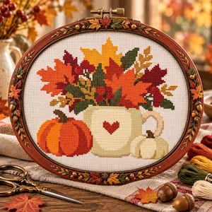 Puede incluir: Un bastidor de bordado ovalado con un diseño de punto de cruz que muestra una taza con un corazón rojo, una calabaza naranja grande, una calabaza blanca pequeña y un ramo de hojas otoñales en tonos rojos, naranjas, amarillos y verdes.