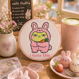 May include: A circular embroidery hoop containing a cross-stitch design of a green frog wearing a pink bunny onesie and holding a carrot. The text "i hate this" is stitched in pink below the frog. The hoop is framed in a light pink plastic border.