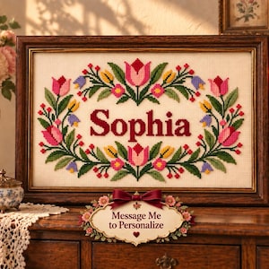 May include: A rectangular cross-stitch embroidery in a dark wood frame featuring the name Sophia in dark red letters, surrounded by a floral border of pink and yellow tulips with green leaves. A small sign below reads Message Me to Personalize.