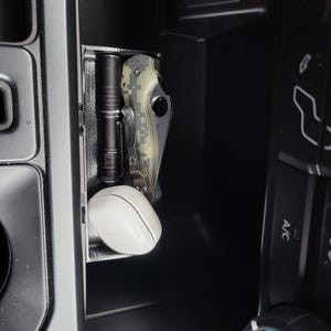 May include: A black and gray flashlight and a camouflage patterned pocket knife are stored in a black plastic organizer in a car's center console.