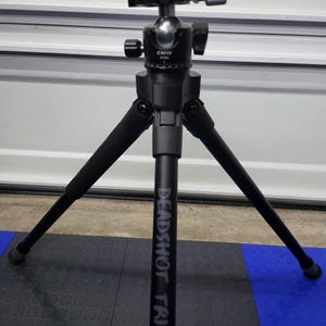 May include: A black Deadshot tripod with three adjustable legs. The tripod has a ball head with a silver knob and black adjustment levers. The text "DEADSHOT TRIPOD" is printed vertically on one of the legs.