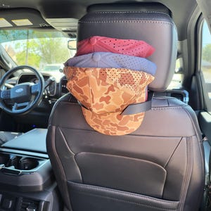 May include: A black plastic hat rack mounted on the back of a vehicle headrest, holding three stacked baseball caps. The top cap is red, the middle is blue, and the bottom cap features a tan and brown camouflage pattern with perforated fabric.