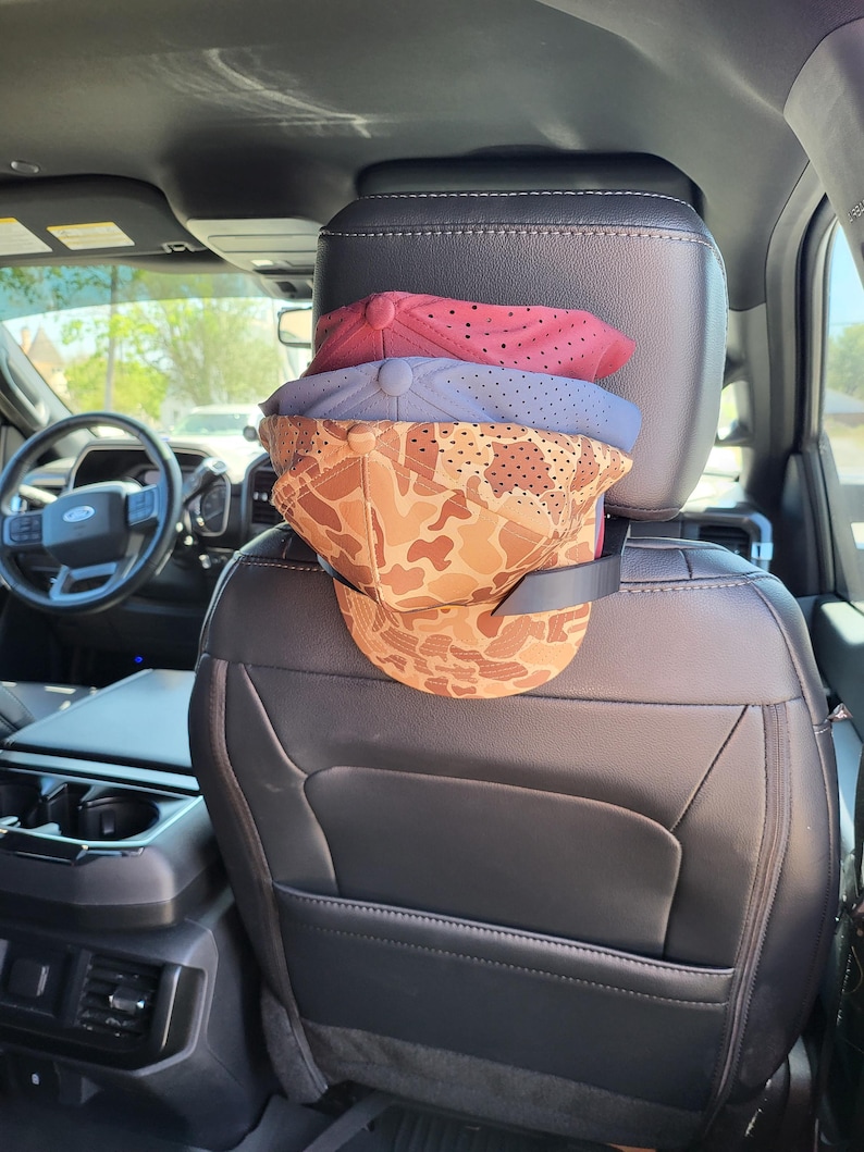 May include: A black plastic hat rack mounted on the back of a vehicle headrest, holding three stacked baseball caps. The top cap is red, the middle is blue, and the bottom cap features a tan and brown camouflage pattern with perforated fabric.