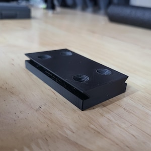 May include: A black plastic bracket with four holes for screws. The bracket has a beveled edge and is designed to be mounted to a surface.