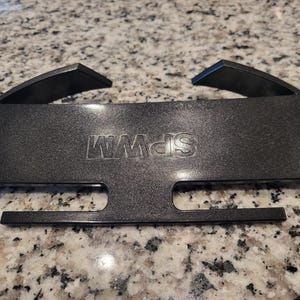 May include: A black plastic rectangular accessory featuring the embossed text SPWM. The object has two angled hooks at the top and two horizontal rectangular cutouts along the bottom edge, with a smooth, slightly textured finish on the surface.