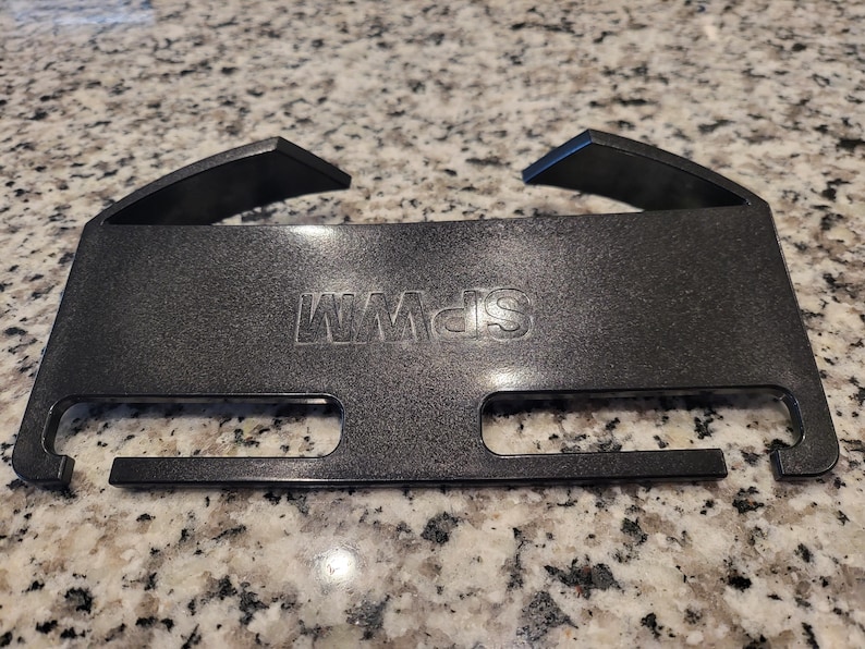 May include: A black plastic rectangular accessory featuring the embossed text SPWM. The object has two angled hooks at the top and two horizontal rectangular cutouts along the bottom edge, with a smooth, slightly textured finish on the surface.