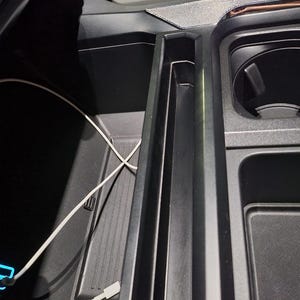 May include: Interior car console with a black finish, featuring a phone charging cable and a cup holder. The console has multiple compartments and a textured surface.