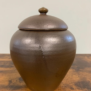 May include: A dark brown ceramic jar with a rounded body and a matching lid featuring a small knob handle. The surface of the pottery has a textured, matte finish with a subtle diagonal line detail running down the side of the vessel.