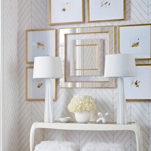 May include: An interior design scene featuring a white console table with two fluffy stools and gold legs. Two white lamps with white shades sit on the table, with framed art and a mirror on the wall. The walls have a white and gold chevron pattern.