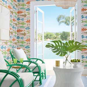 May include: Two green wicker chairs with white cushions sit on a blue and white striped rug in front of a large window with a view of a swimming pool and palm trees. A white round table with a vase of green leaves and a bowl of limes sits between the chairs.