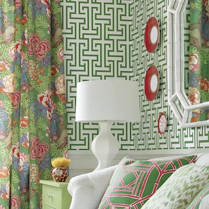 May include: A bright living room scene with green floral curtains, a white sofa, and patterned throw pillows. A white lamp sits on a green side table. The wall features a green geometric pattern and decorative red plates.