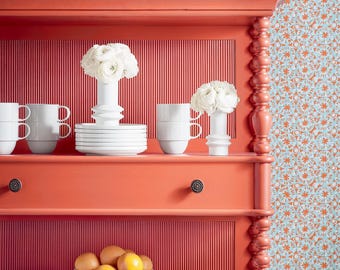 May include: A coral red hutch with two shelves, a drawer, and a decorative trim. The hutch is filled with white dishes, mugs, and vases with white flowers. There are also oranges in a white bowl on the bottom shelf.