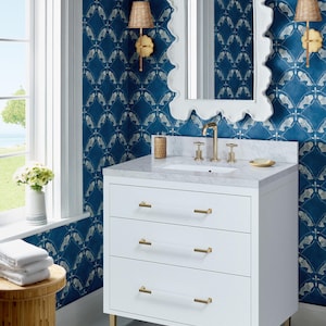 May include: White bathroom vanity with three drawers and gold hardware. The vanity has a white countertop with a white porcelain sink and gold faucets. The vanity is in front of a blue and white patterned wallpaper with a white framed mirror.