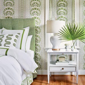 May include: A bedroom scene featuring a white nightstand with a lamp, books, and a vase of flowers. The bed has white linens with green trim and decorative pillows. The headboard and wallpaper have a green and white floral pattern.