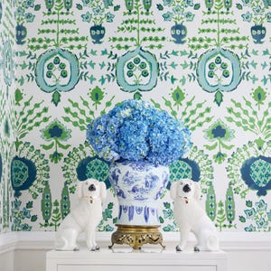May include: A white console table with a single drawer and a gold cross-shaped pull. The table is decorated with a blue and white porcelain vase filled with blue hydrangeas and two white porcelain dog figurines.