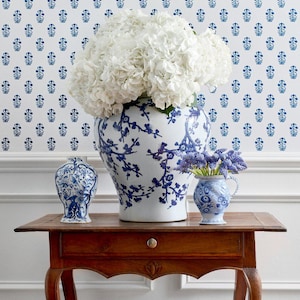 May include: A wooden table displays three blue and white ceramic vases, each with a unique design. The largest vase holds white flowers, while the smaller ones contain blue flowers. The background features a white wall with a blue floral pattern.