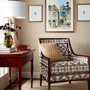 May include: A brown armchair with a colorful geometric patterned upholstery and a brown throw pillow. The chair is in a room with a striped wallpaper, a wooden table, and a lamp with a white lampshade.