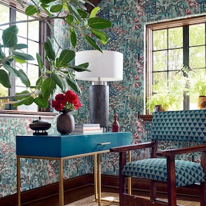 May include: A teal blue desk with gold metal legs and a drawer. The desk is in a room with a patterned wallpaper, a wooden chair with a teal blue and green geometric pattern, and a lamp with a white shade.