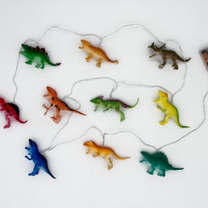 Pode incluir: Uma sequência de luzes de dinossauros coloridos. A sequência apresenta várias figuras de dinossauros em vermelho, verde, laranja, amarelo e azul. As luzes são alimentadas por um pacote de baterias.