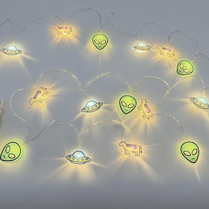 Op de afbeelding: Slinger met lichtjes met verlichte alienkoppen, UFO's en koeienontwerpen. De lampjes geven een warme gloed. De batterijhouder is links zichtbaar. Perfect voor een speelse decoratie in elke ruimte.