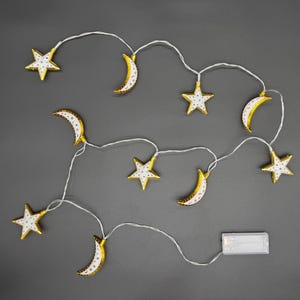 Puede incluir: Una cadena de luces decorativas con estrellas y lunas crecientes doradas con detalles blancos. Las luces están ensartadas en un cable blanco y funcionan con una batería. Fondo gris.