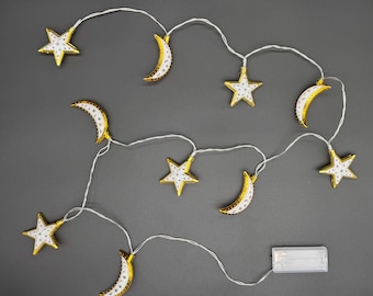 May include: A string of decorative lights featuring gold stars and crescent moons with white detailing. The lights are strung on a white wire and powered by a battery pack. The lights are set against a grey background.