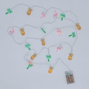 May include: String of decorative lights featuring pineapple, flamingo, and palm tree shapes. The lights are in various colors, including yellow, pink, and green, and are strung on a white wire. A battery pack is visible at the end of the string.